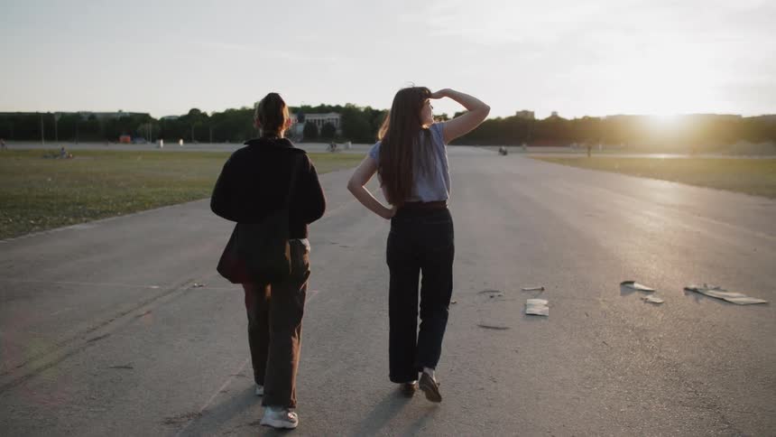 Two women walk side by side on an open paved path as the sun sets in the distance