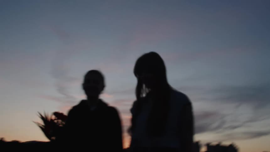 Two female friends one holding flowers walk outdoors hand in hand as darkness slowly sets in