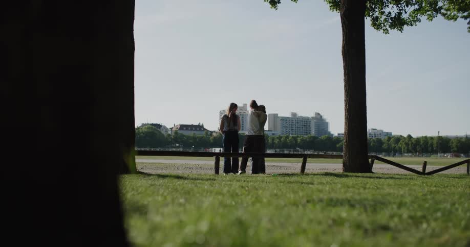 In a park on a sunny day three young people are sitting and talking on a bench with a city skyline in the background