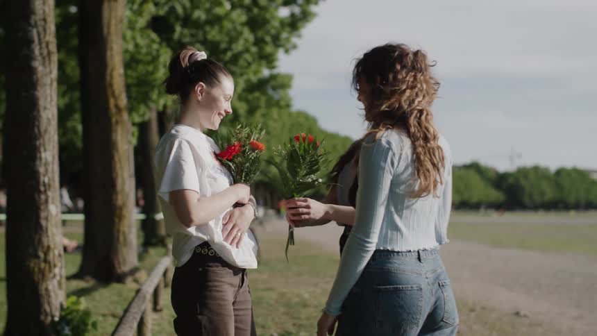 Two women delivering flowers to a friend while standing in a park surrounded by trees