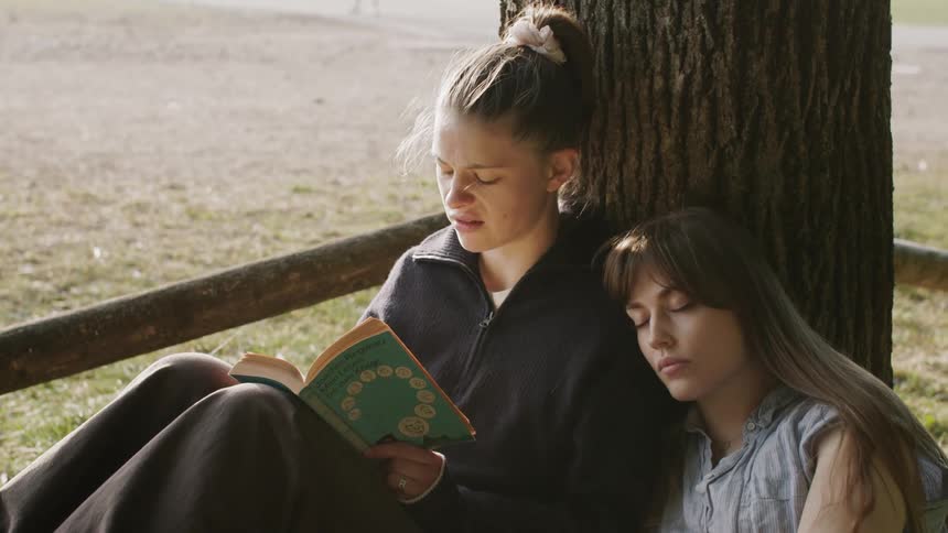 Two young women sit under a tree as one reads a book to the other who looks sleepy