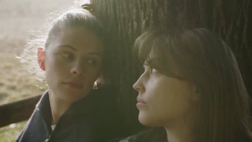 Two teenage girls sit on the floor resting their backs against a tree with their eyes closed suddenly they open their eyes and stare at each other at midday