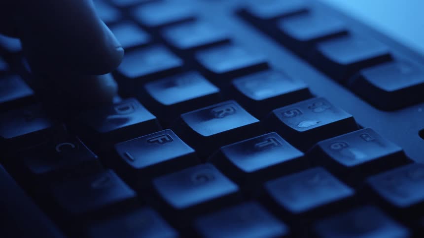 Close up of a desktop computer keyboard as a person types with it in a blue lit room