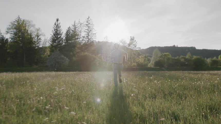 Back view of a male running in a field towards a treeline he is wearing jeands and a long sleeve shirt his hair is bouncing as he runs the sun is casting a glowing light on the male