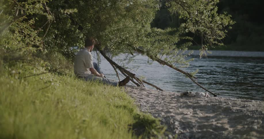 Man sitting under a tree by a riverbank using a smartphone