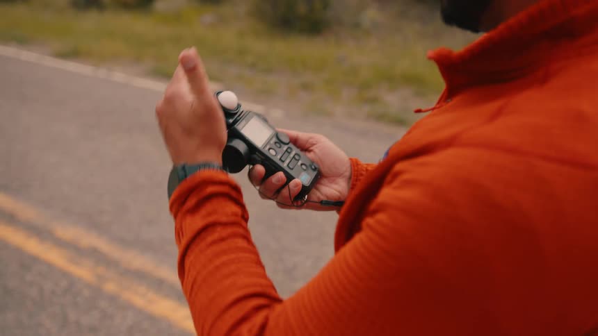 A man wearing an orange shirt stands in the middle of the road at midday holding a professionalgrade light meter