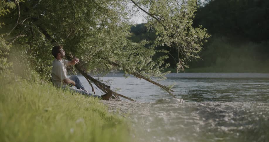 A person sits alone on a grassy riverbank gazing across the calm water framed by dense trees and soft daylight