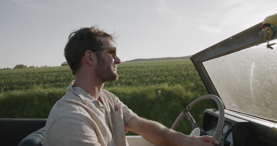 A man drives a vintage convertible along a narrow rural road flanked by bright fields under a clear sky