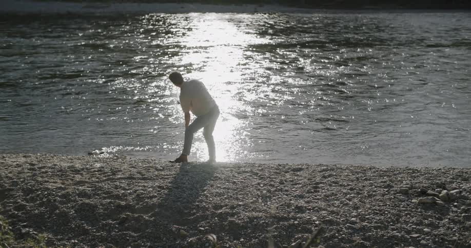 A man on the bank of a lake picks up a rock from the ground and skips it across the water at midday with sunlight reflecting off the lake