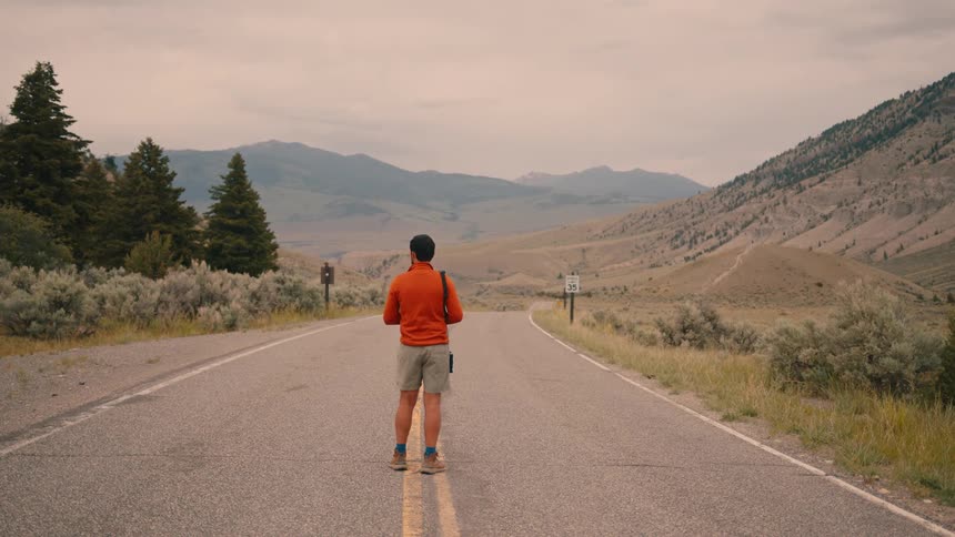 A man stands alone on a rural mountain road looking ahead at the scenic landscape