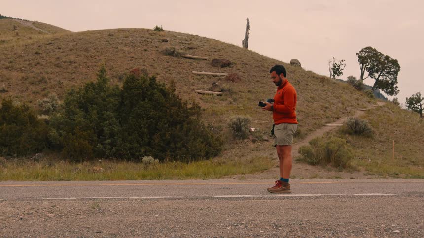 A young man walks in the middle of the road surrounded by hills and grassland then takes out a camera and starts taking photos at midday