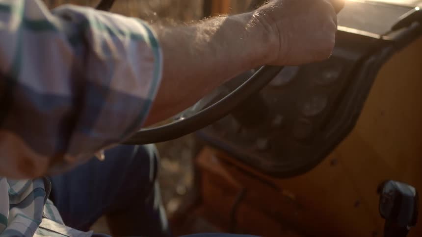 A closeup of a persons hands gripping a steering wheel likely of a tractor or other farm machinery the golden light in the background suggests it might be sunrise or sunset and hints at an agricultural setting