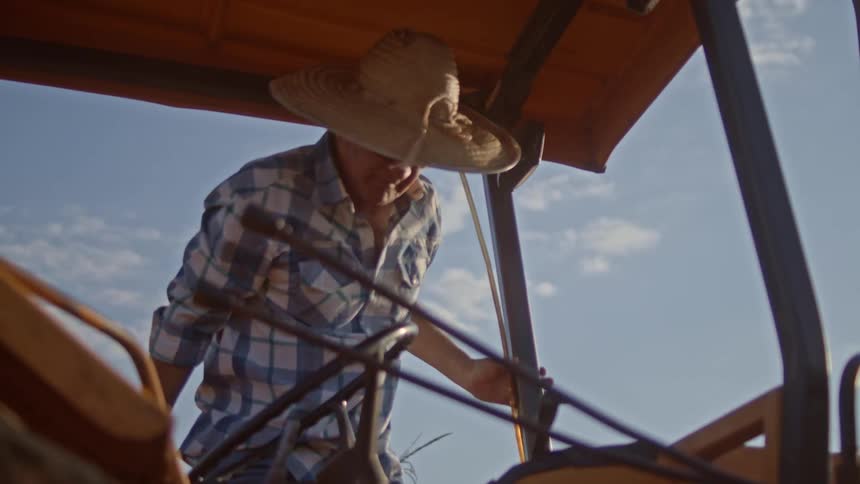 A man in a straw hat and blue and white checkered shirt boards a tractor from a low angle beneath a clear sky