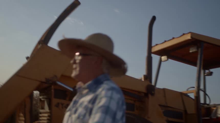 A man wearing a straw hat and plaid shirt is stepping out of a yellow tractor in a dry harvested field under a bright clear sky