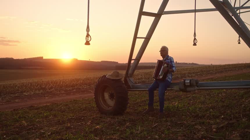 An older man plays accordion beside irrigation gear in a field at sunset