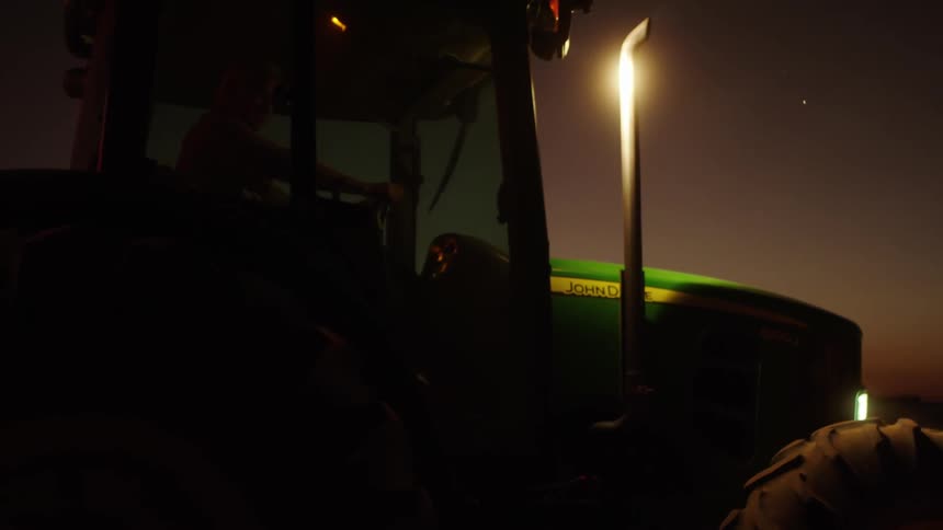 A male farmer drives a green farm tractor at night