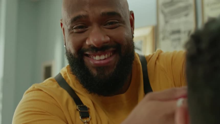 A man is barbering as the camera pans to him he smiles at the camera looking away from the person he is working on
