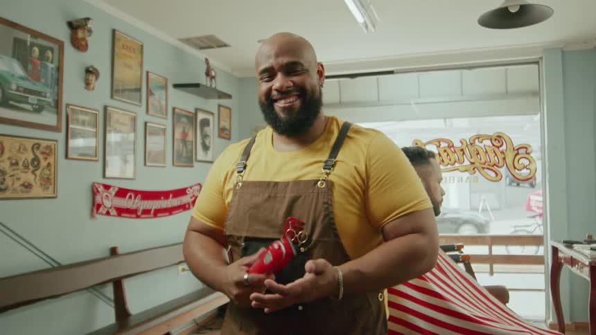 A smiling barber taps an electric razor in his hands while his draped client waits