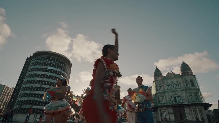 A performer in a vibrant red and yellow costume jumps in midair with a yellow umbrella surrounded by other performers in a city square