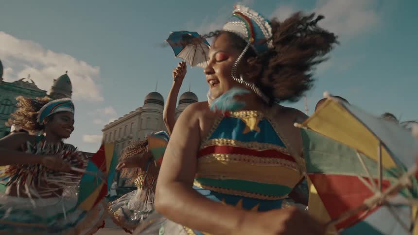 A group of people in native attire dance in the middle of the street holding small umbrellas