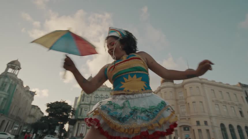 A woman in colorful traditional clothing dances joyfully in a historic city square waving a multicolored flag