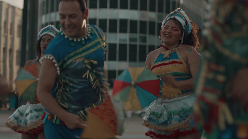 Dancers in vibrant traditional costumes joyfully perform in the street with one woman holding a colorful umbrella and others moving rhythmically around her during a festive celebration