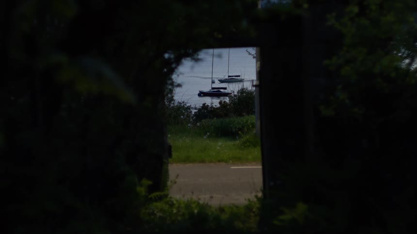 A sailboat is visible in the distance through a shaded leafy frame of trees and a stone archway
