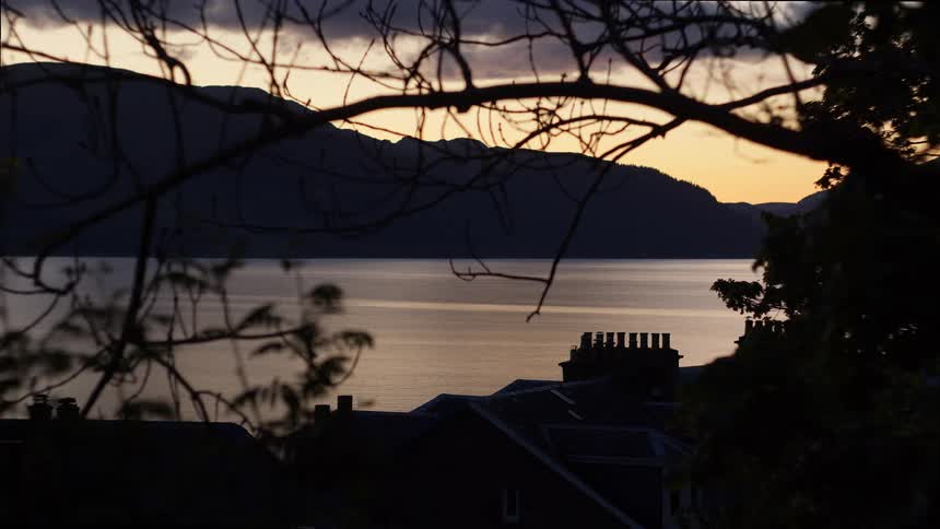 Silhouetted trees and rooftops frame a tranquil lake and mountain at sunset