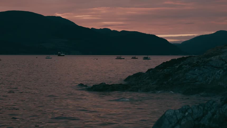 Boats float on a calm coastal bay at sunset with silhouetted mountains in the background