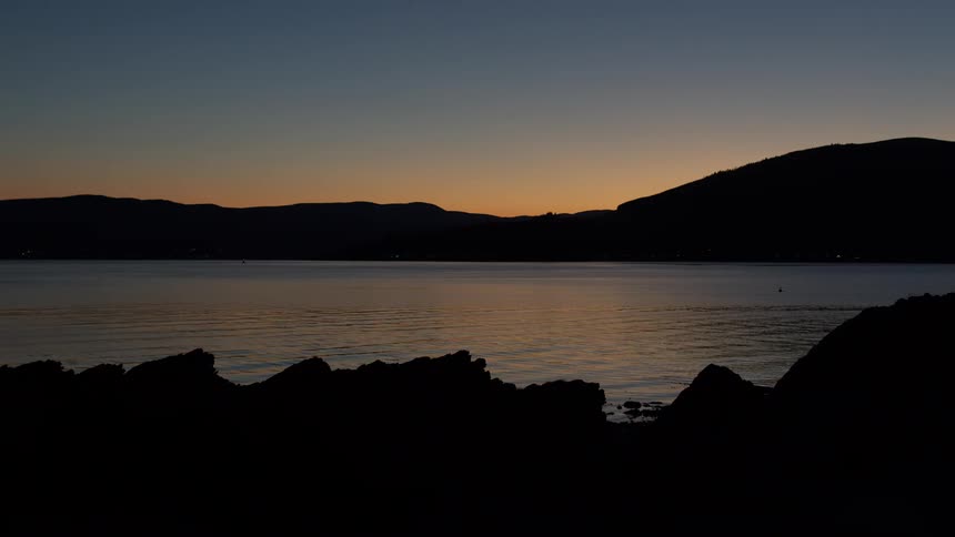 A calm coastal scene at dusk with mountains silhouetted against a fading orange sky