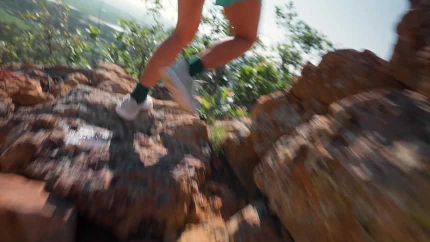 A person wearing teal shorts and trail shoes is running along a rocky sunlit dirt path on a hillside surrounded by rugged terrain and greenery