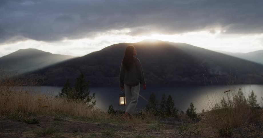 A woman stands holding a lantern gazing at a mountain and lake during sunset