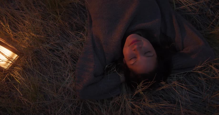 A woman lies in dry grass with hands behind her head gazing calmly upward near a warm light