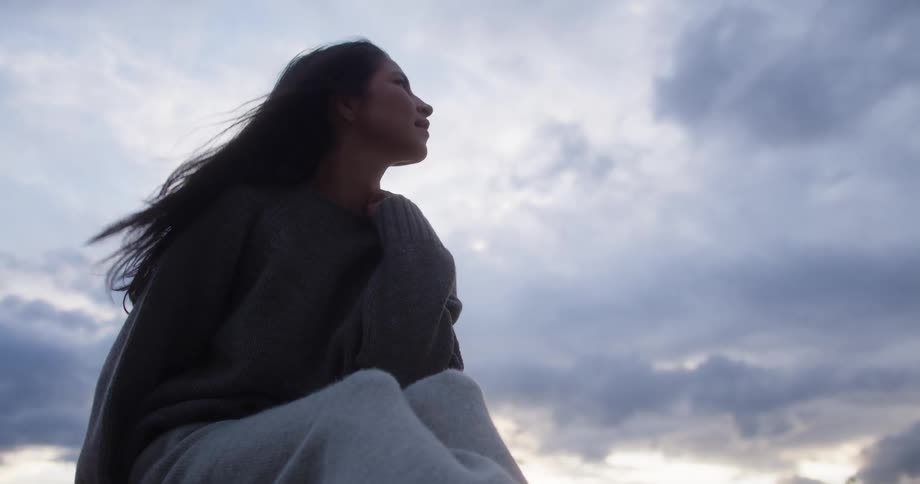 A woman wrapped in a sweater gazes thoughtfully at a cloudy sky