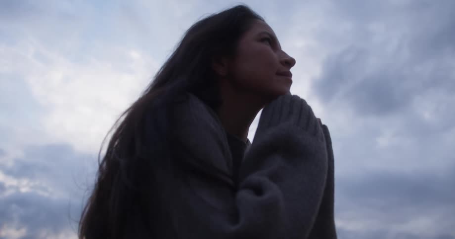 A young woman in a sweater sits on a rock while outdoors under a cloudy sky