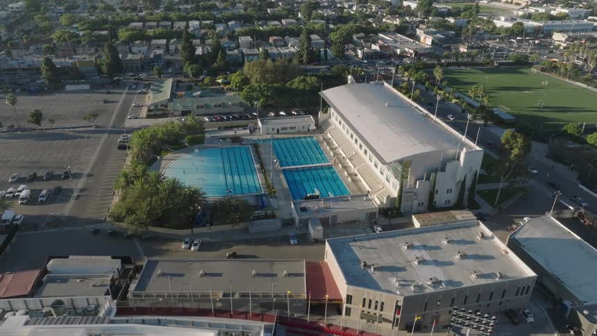 An aerial view shows Los Angeles Swimming Stadium with two large Olympic pools beside a modern building surrounded by parking lots and city streets in Los Angeles