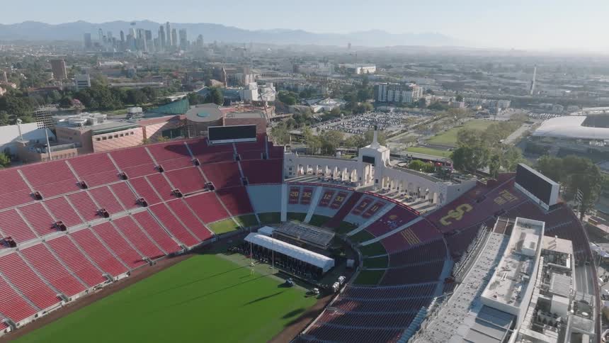 Aerial view of an empty Los Angeles Memorial Coliseum stadium with a city skyline and mountains in the background