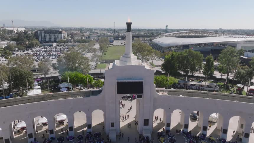 Los Angeles Memorial Coliseum with red and white stands displays historical figures next to a monumental portico with columns and Olympic Torch caldron