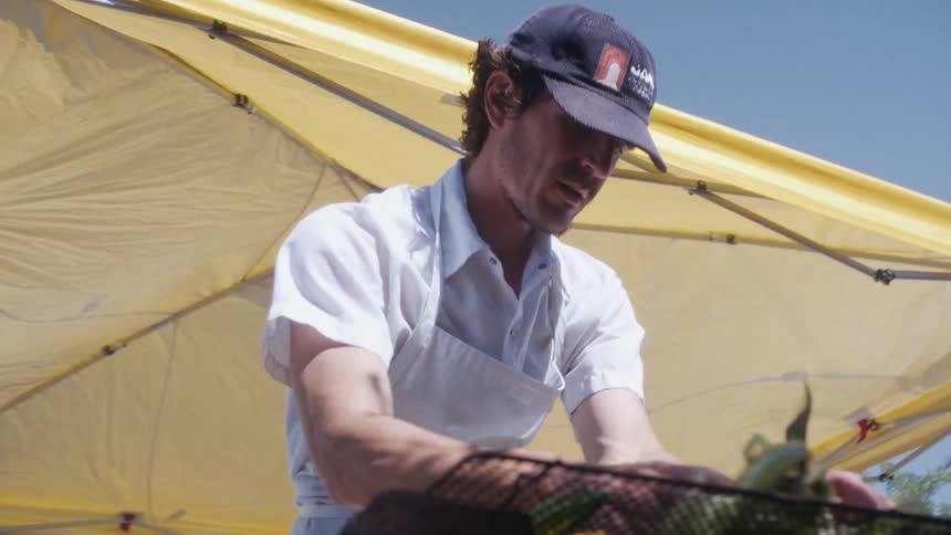 A chef prepares food on a grill under a canopy outdoors at midday