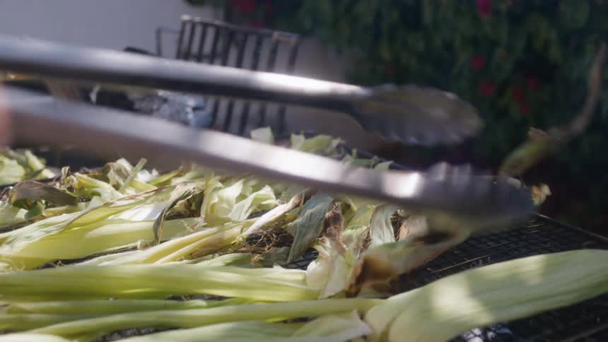 A person uses tongs to grill corn in husks over hot coals with roasted vegetables visible on a tray in the background