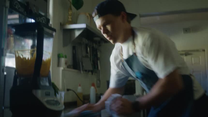 Man in cap and apron cleaning sink in restaurant kitchen with blender in action nearby
