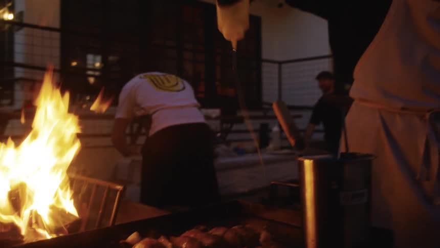 A man stands beside a grill pouring oil on top of it outdoors at sundown while others behind him make preparations