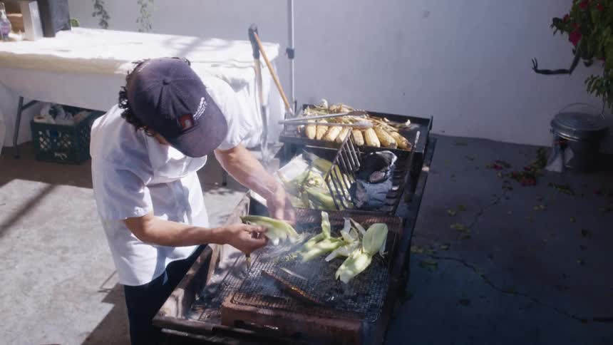Young male chef grills corn and its husk at an outdoor grill during daytime