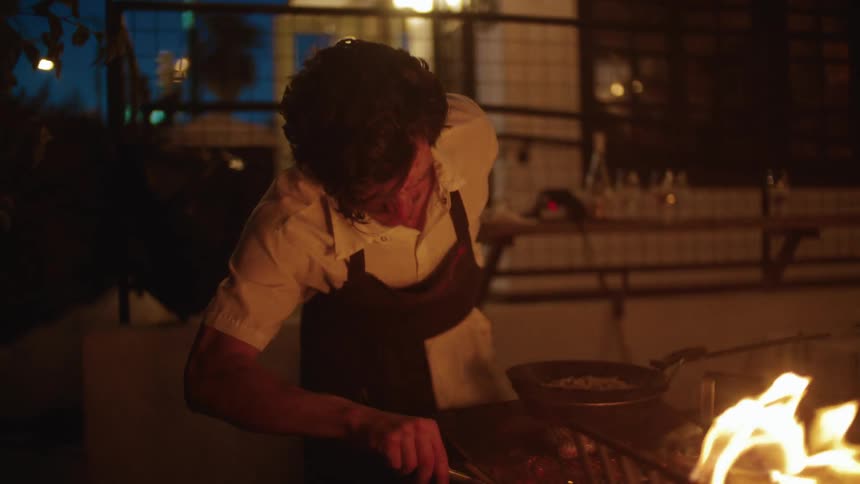 A chef carefully tends to food on a flaming grill in a warmly lit outdoor kitchen at night