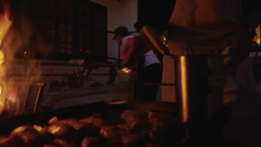 Chef grills meat over an open flame in a dimly lit kitchen