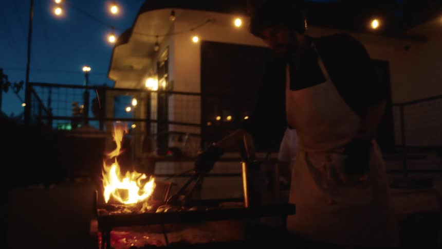 A chef grills food over open flames at night under string lights