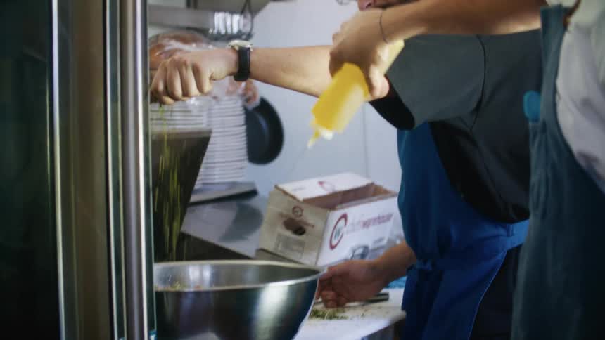Two people prepare food in a kitchen one holding a bottle over a mixing bowl