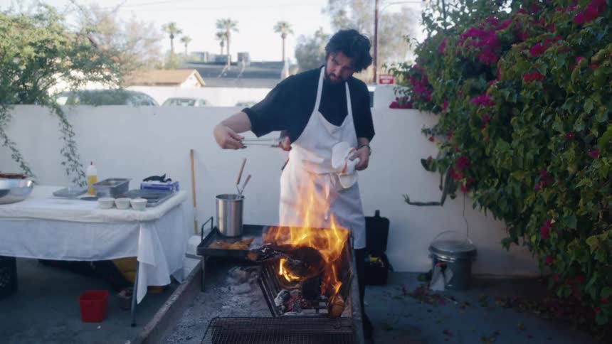 Chef grills over open flames outdoors using long tool