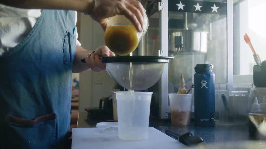 A person strains juice through a sieve into a plastic container in a kitchen setting