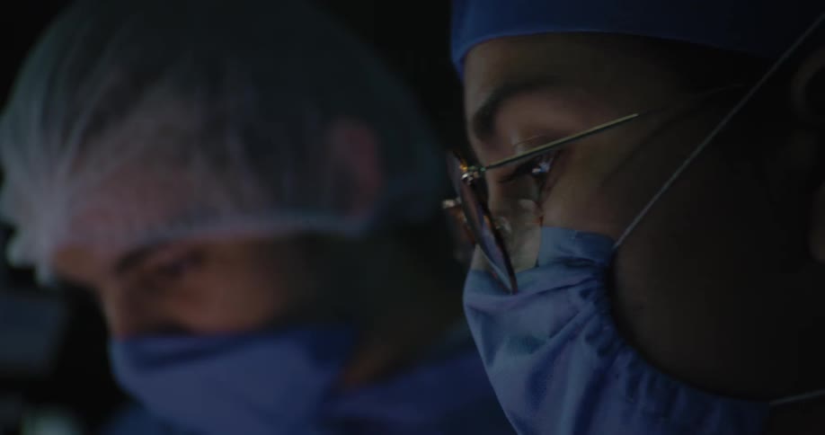 Closeup of the faces of two medical professionals both wearing surgical masks blue scrubs and caps one wears glasses and the background is dim
